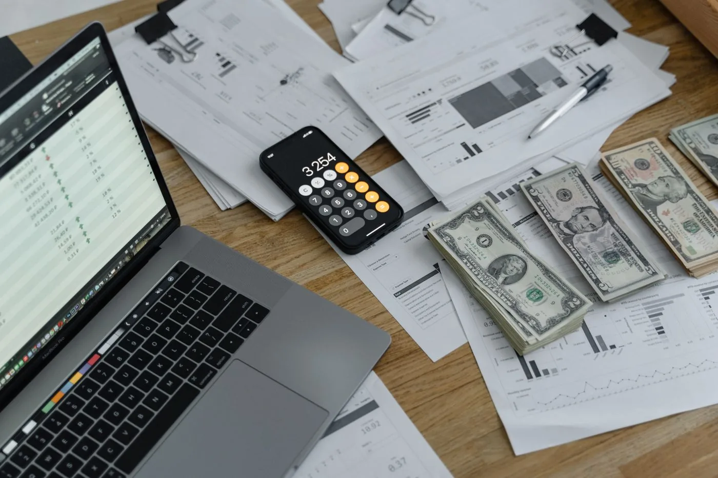 No copyright restriction A wooden desk with a laptop, stacks of United States bills, a phone calculator, and papers.