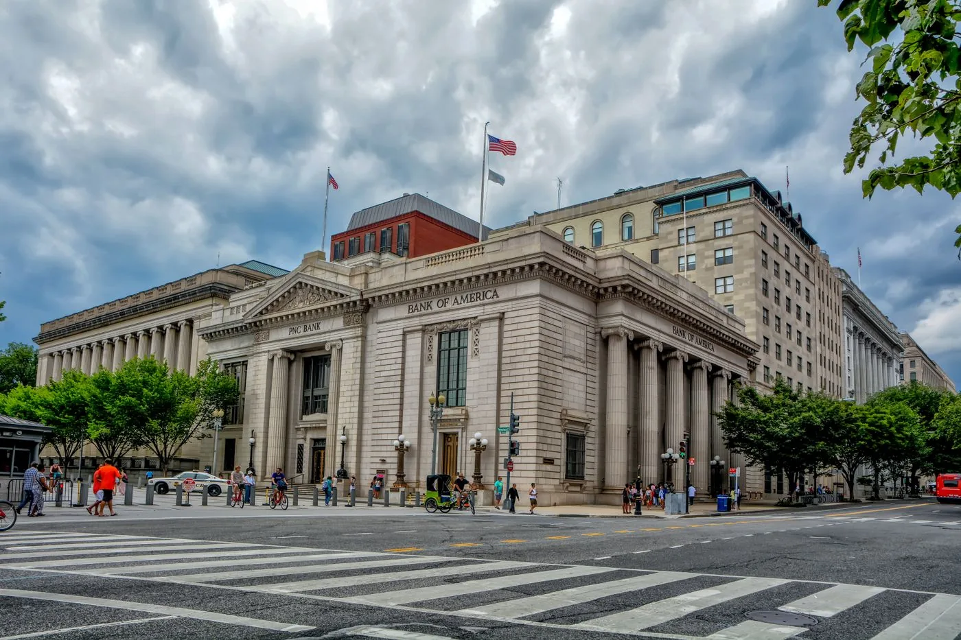 A stone bank building on a city corner with columns, flags, and a crosswalk.